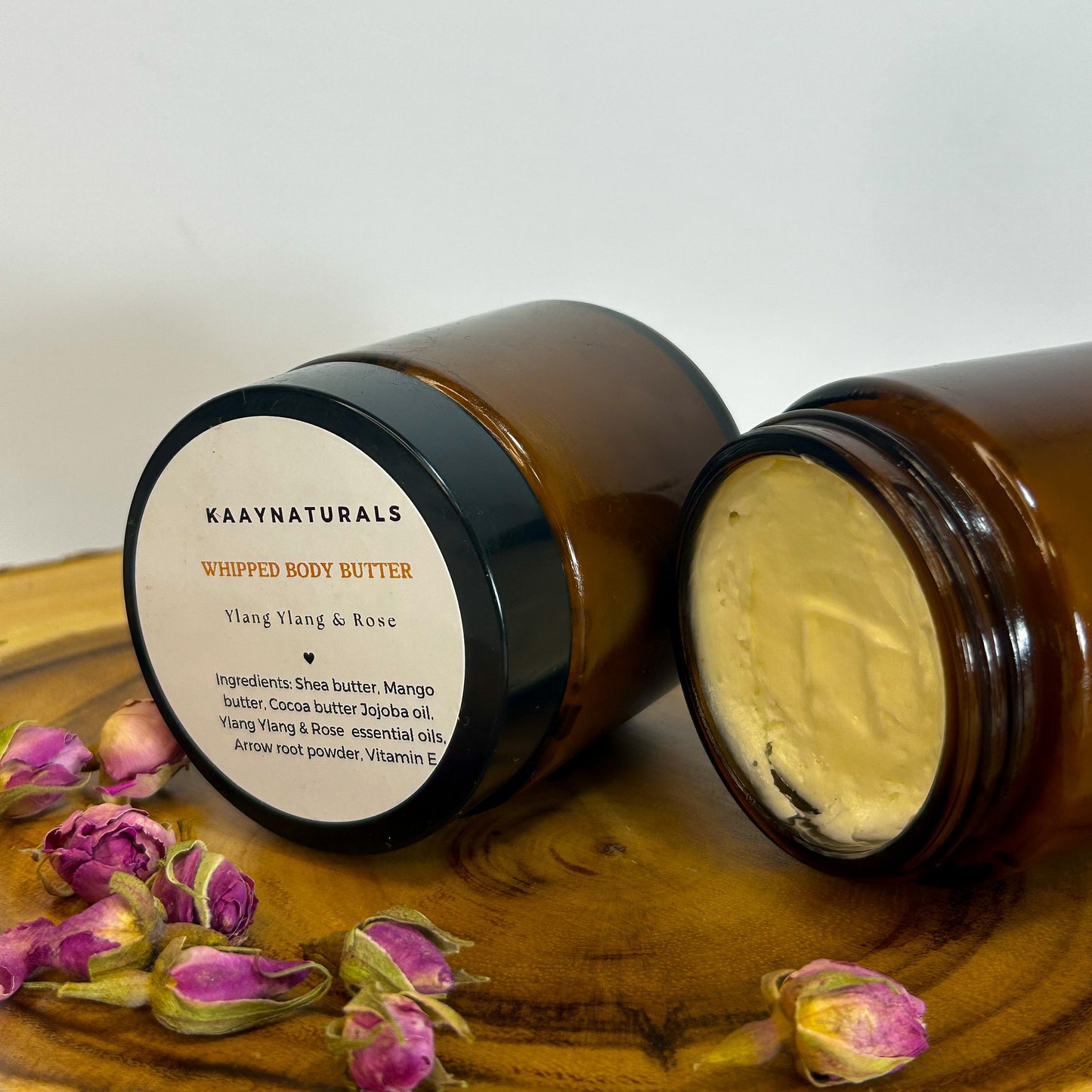 Brown jar of KAYANATURALS whipped body butter with a visible label on a wooden surface with pink flowers.